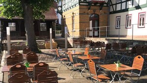 An outdoor seating area with wooden tables and chairs, a tree, and a building with a traditional facade in the background.