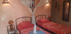 Habitación con dos camas, cada una con cubrecama y almohadas rojas, una mosquitera sobre las camas, una mesita con una lámpara y un cuadro en la pared.