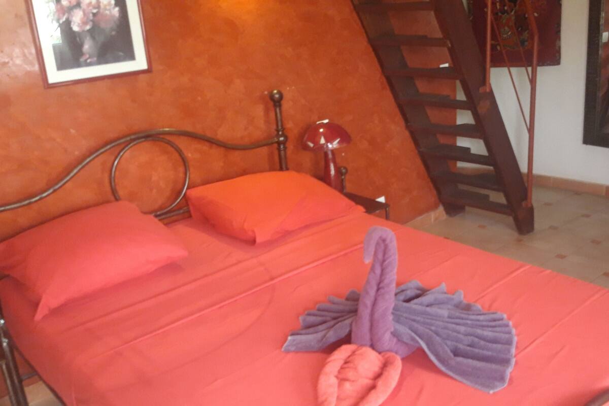 A bed with a red bedspread, a purple towel, and a pink pillow in a room with a wooden ladder and a framed picture on the wall.