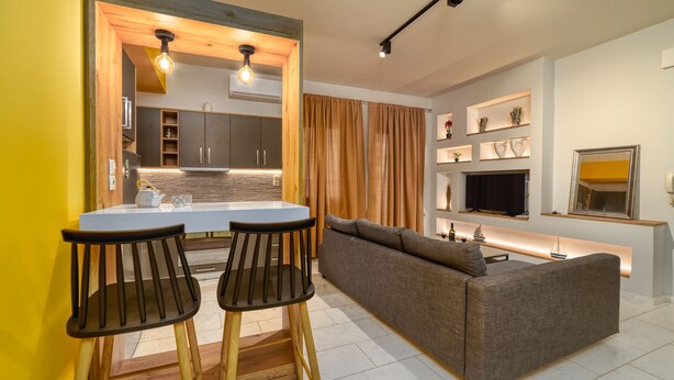 A modern kitchen with a central island, two bar stools, a sofa, and a TV on a stand. - Amoudara Suites (Malevizi)