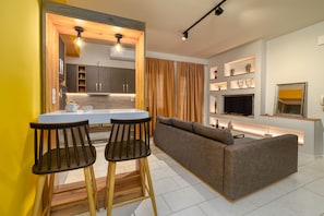 A modern kitchen with a central island, two bar stools, a sofa, and a TV on a stand. - Amoudara Suites (Malevizi)