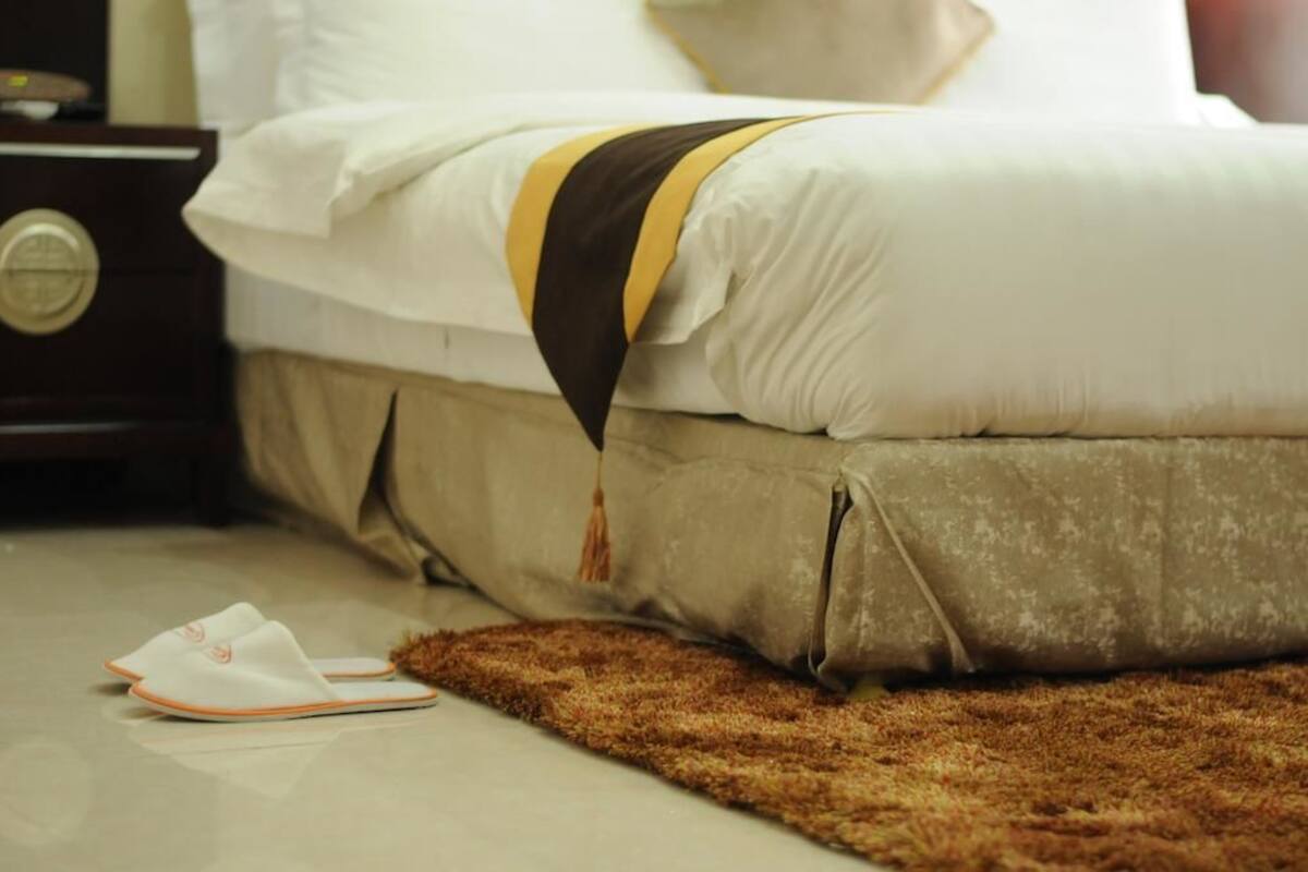 Un lit recouvert d’une couverture blanche et marron, un tapis marron et une paire de chaussons posés sur le sol.
