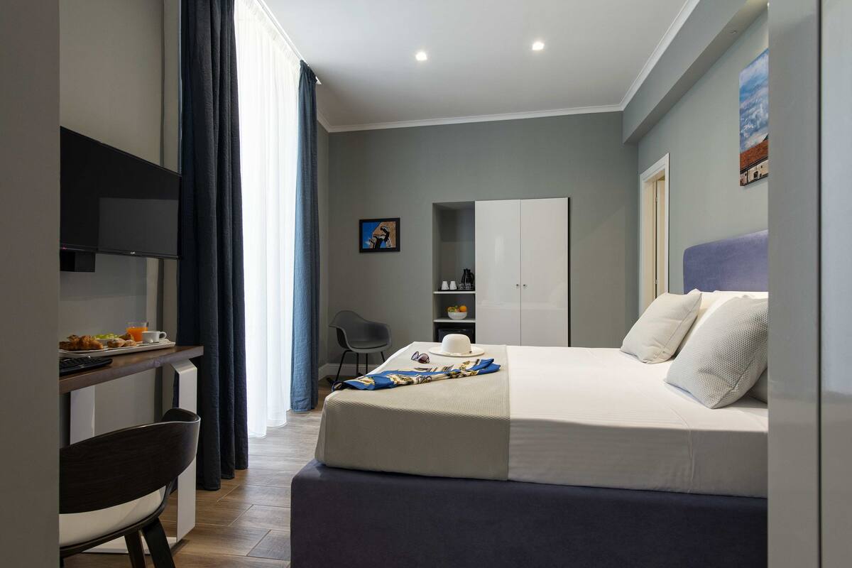 A modern hotel room with a bed, a desk, a chair, a wardrobe, and a television.