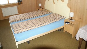A room with two beds, wooden headboards, a small bedside table with a lamp, a radiator, and a window with curtains.