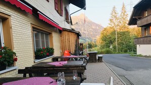 An outdoor dining area with wooden tables and benches, potted plants, and a mountain view in the background.
