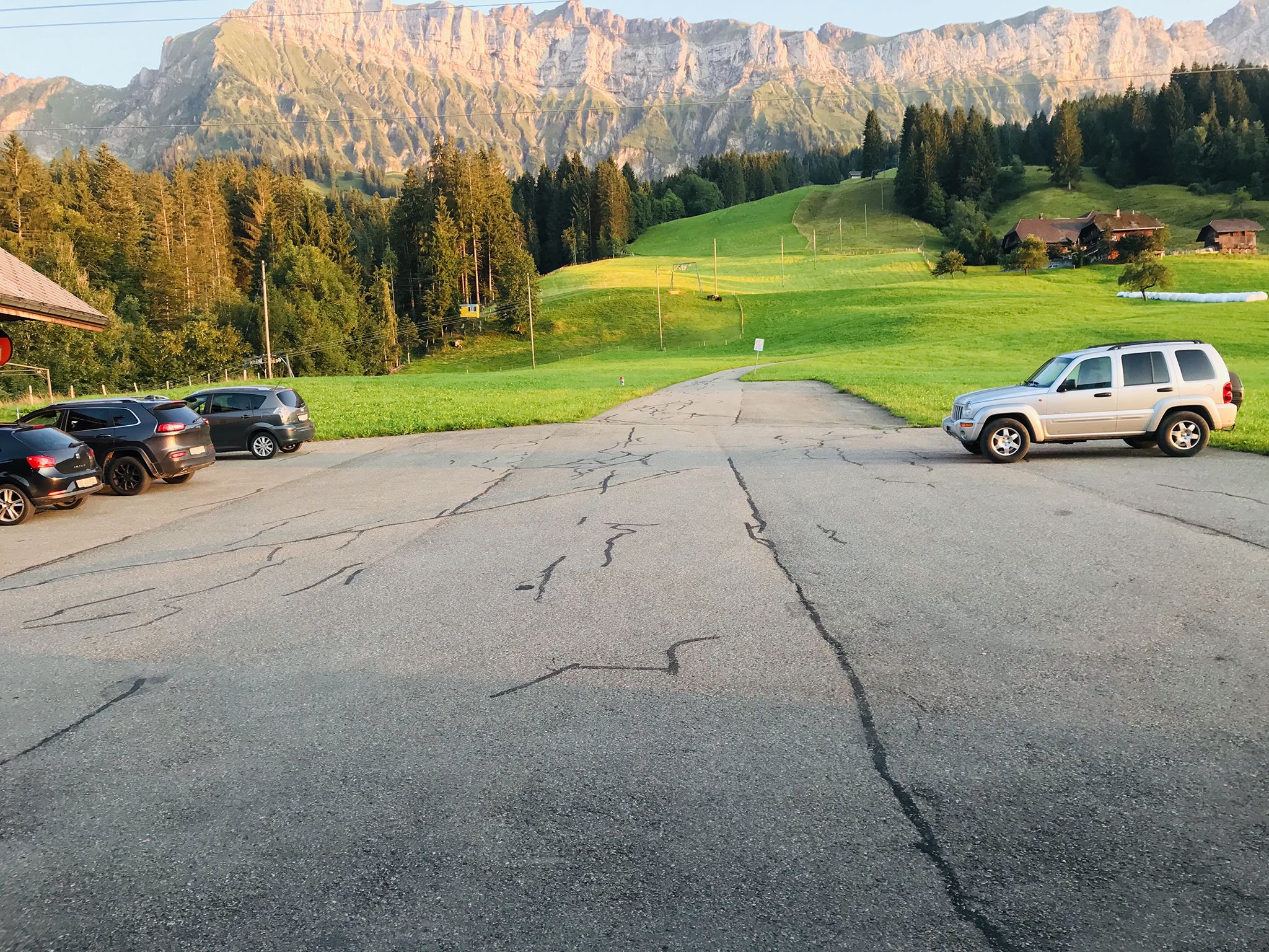 Ein Parkplatz mit mehreren Autos, einem weißen SUV und Bergelandschaft im Hintergrund.