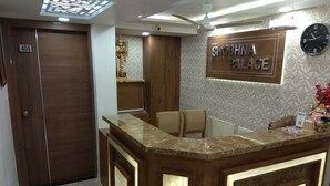 Un comptoir d’accueil avec une enseigne indiquant « SHOBHNA PALACE » et une horloge. On y trouve une porte portant le numéro « 404 » et un ventilateur de plafond.