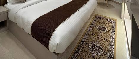 A modern hotel bedroom with a large bed, a headboard, bedside tables, a desk, a chair, a TV, and a bathroom.