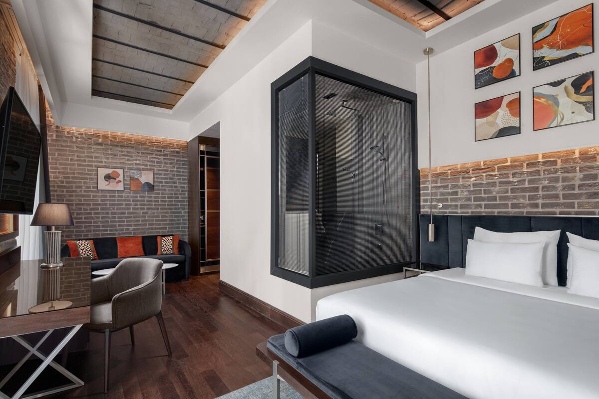 A modern hotel room with a large bed, a desk with a chair, a sofa, and artwork on the walls.