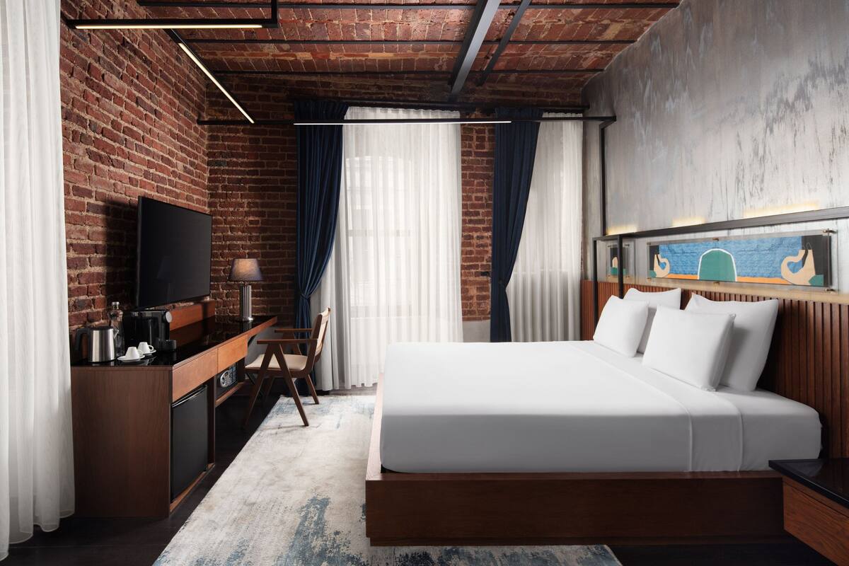 A modern hotel room with a brick wall, a flat-screen TV, a wooden desk with a coffee maker, a bed with white bedding, and a painting above the bed.