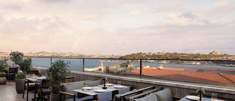 An outdoor dining area with tables and chairs, overlooking a waterfront cityscape.