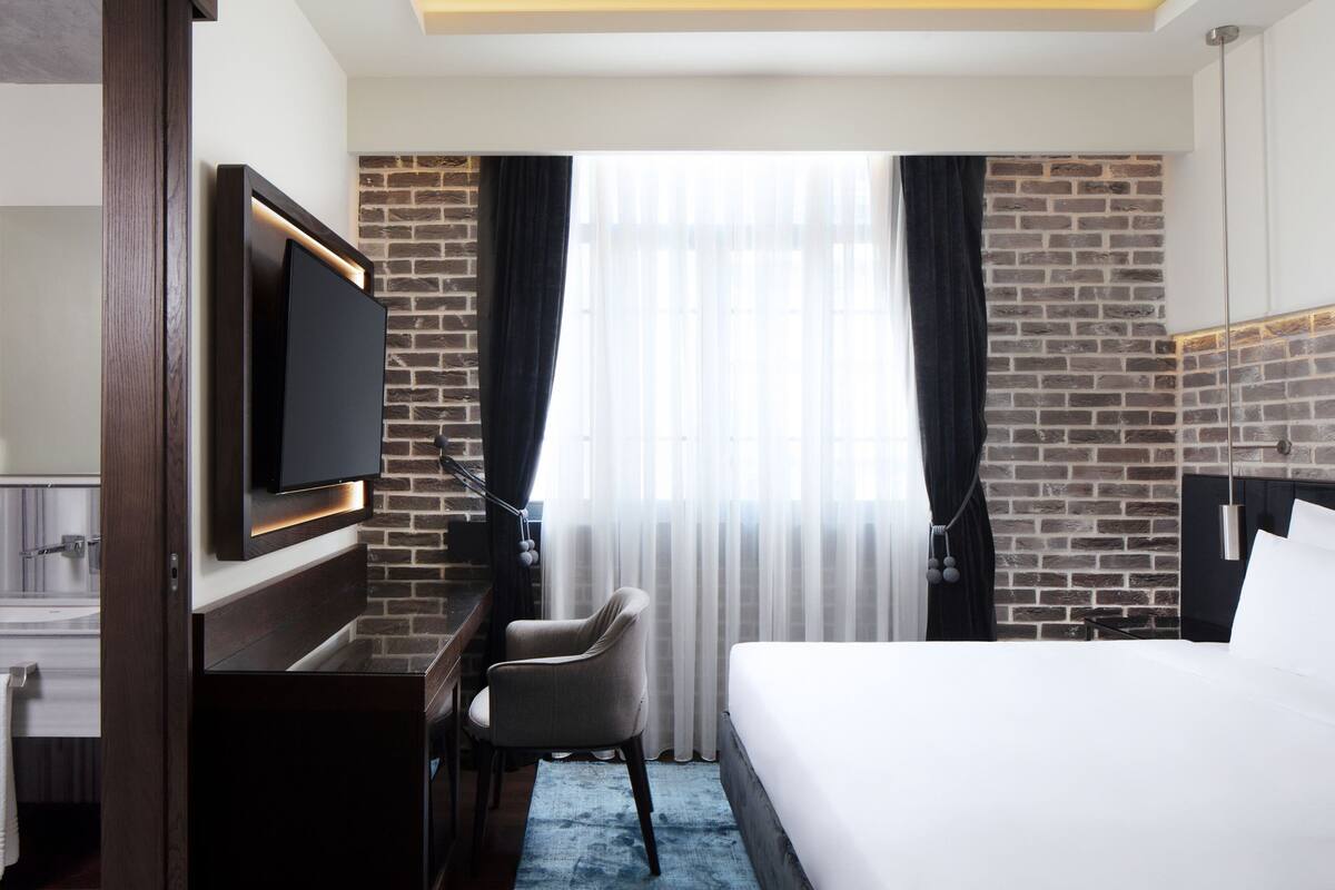 A hotel room with a brick wall, a flat-screen TV mounted on the wall, a bed, a desk with a chair, and a large window with sheer curtains.