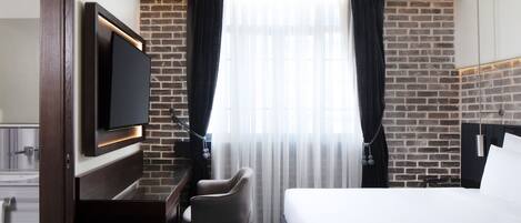 Habitación de hotel con pared de ladrillo, televisor de pantalla plana montado en la pared, cama, escritorio con silla y un amplio ventanal con cortinas de encaje.