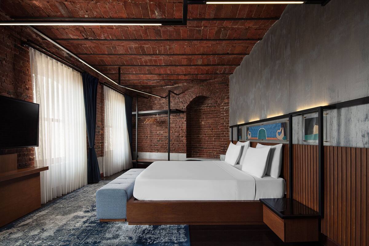 A modern hotel room with a brick ceiling, a large bed, a flat-screen TV, and a large window with sheer curtains.