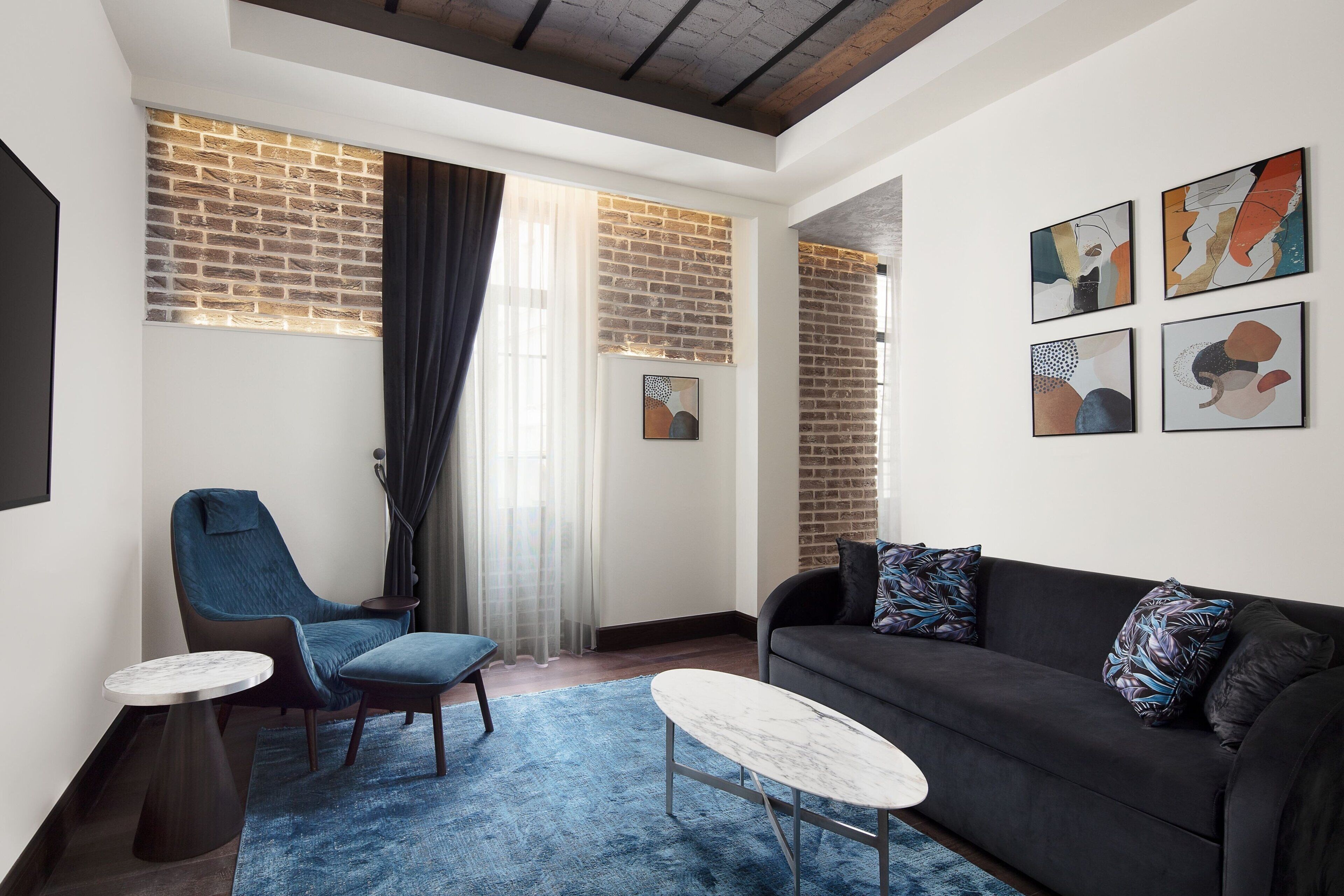 Una sala moderna con pared de ladrillo decorativa, un sofá negro, un sillón azul, una mesita de centro redonda y arte abstracto en la pared.