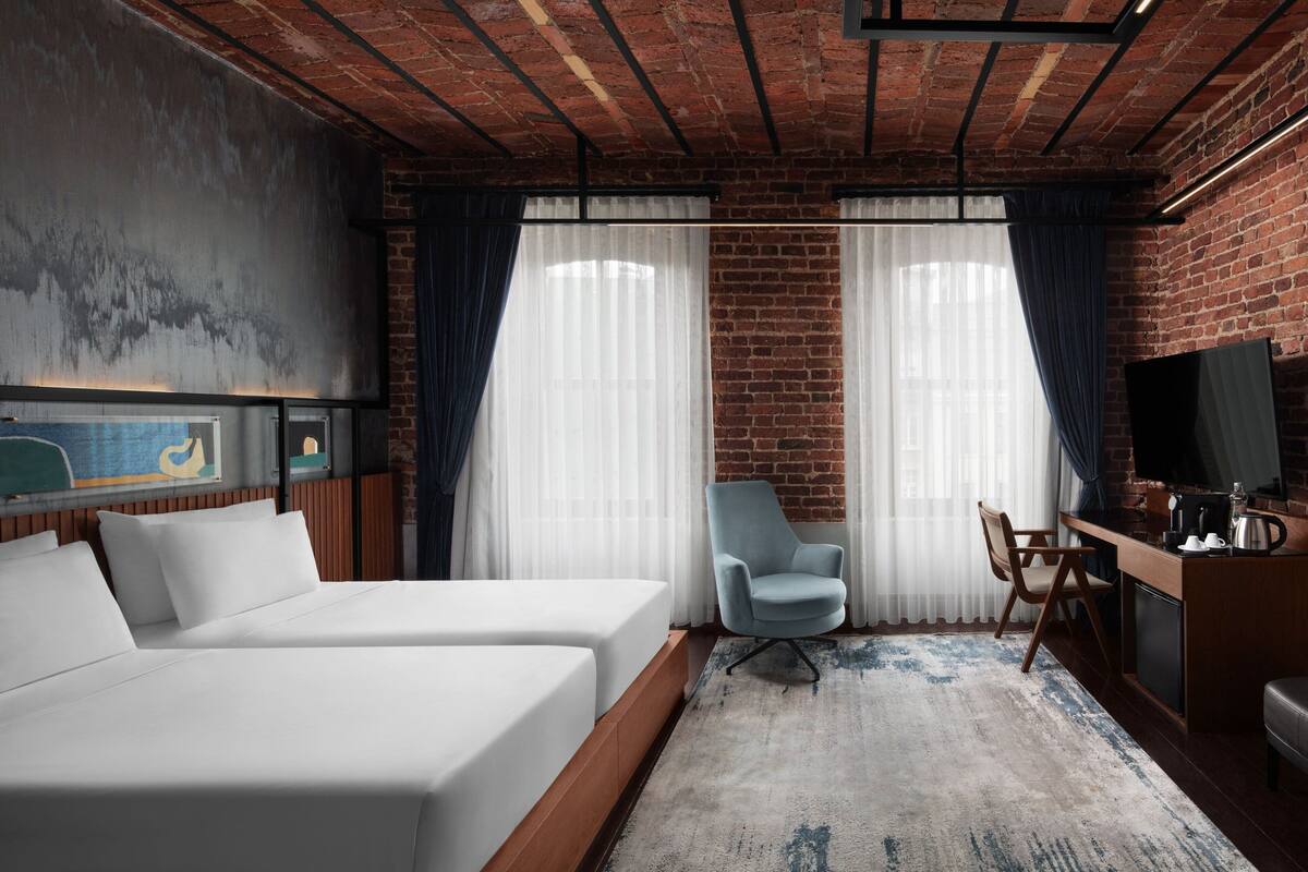 A hotel room with a brick wall, two beds, a desk with a chair, a television, and a large window with curtains.