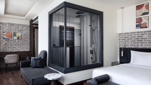 Habitació d'hotel moderna amb un llit gran, una zona de seients amb un sofà i una butaca, una taula rodona petita i un espai de banyera amb vidre.