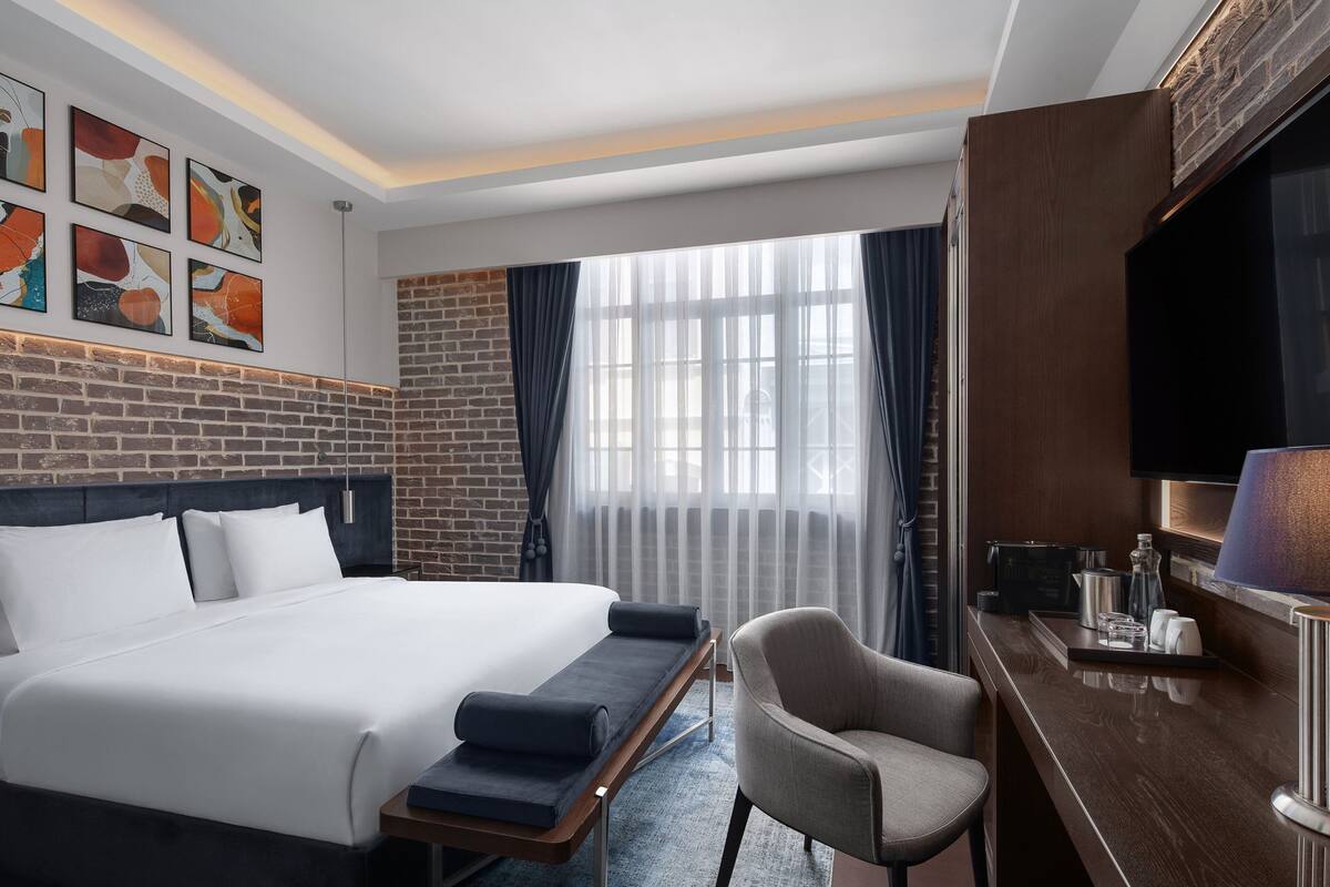 A hotel room with a large bed, a desk with a lamp, a chair, and artwork on the wall.