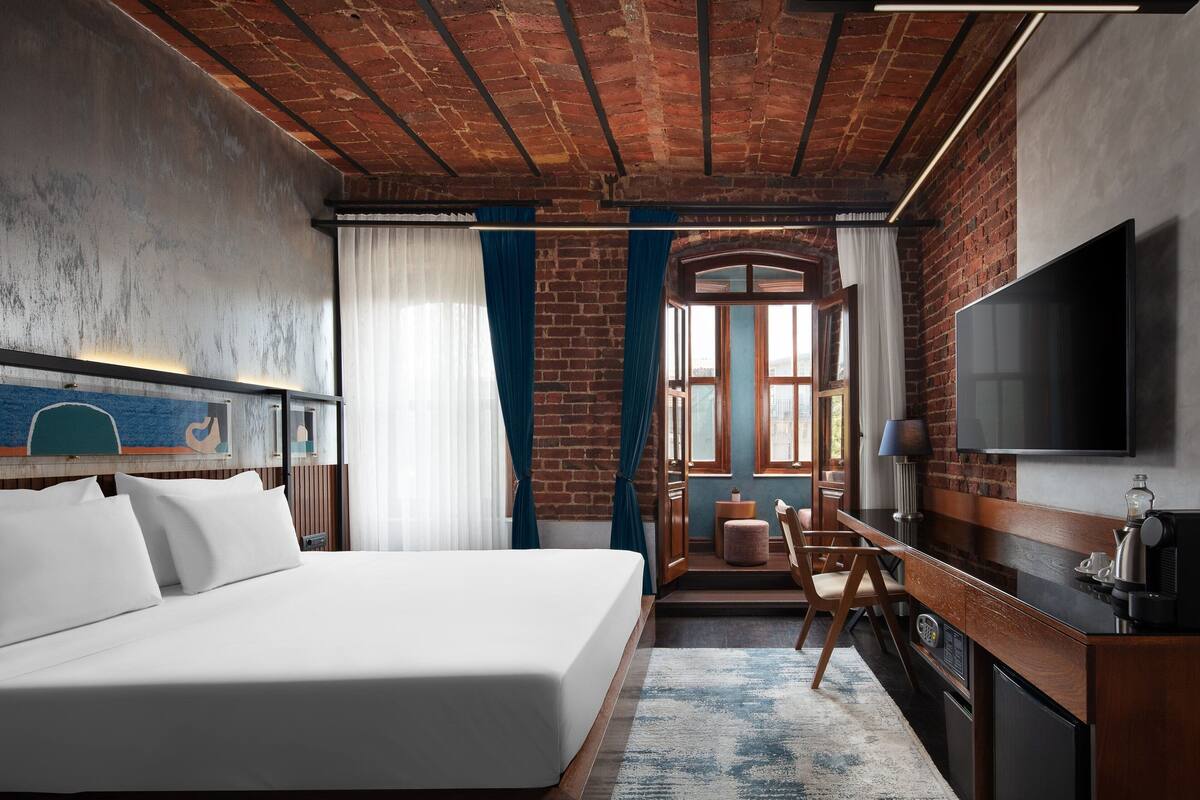 A hotel room with a large bed, a TV, a desk with a chair, and a brick wall.