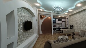 A modern living room with a sofa, a coffee table, and a TV mounted on the wall. There is a kitchen area with a sink and cabinets visible in the background.