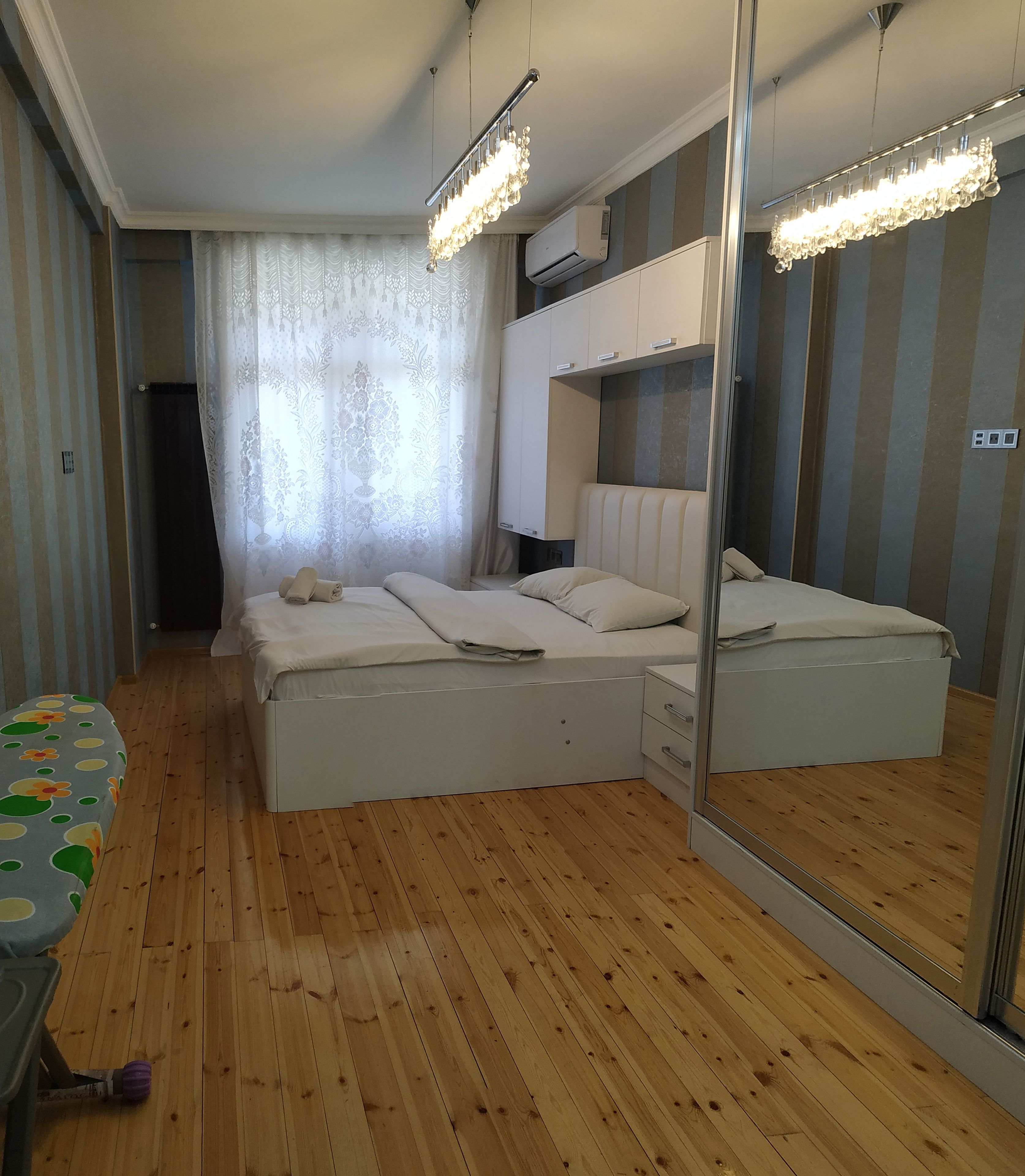 Un dormitorio con suelo de madera, cama con ropa blanca, un espejo grande y una lámpara en el techo.