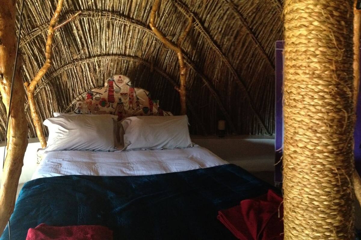 A traditional room with a thatched roof, a bed with a white and blue bedspread, a small table with a vase, and a wicker chair.