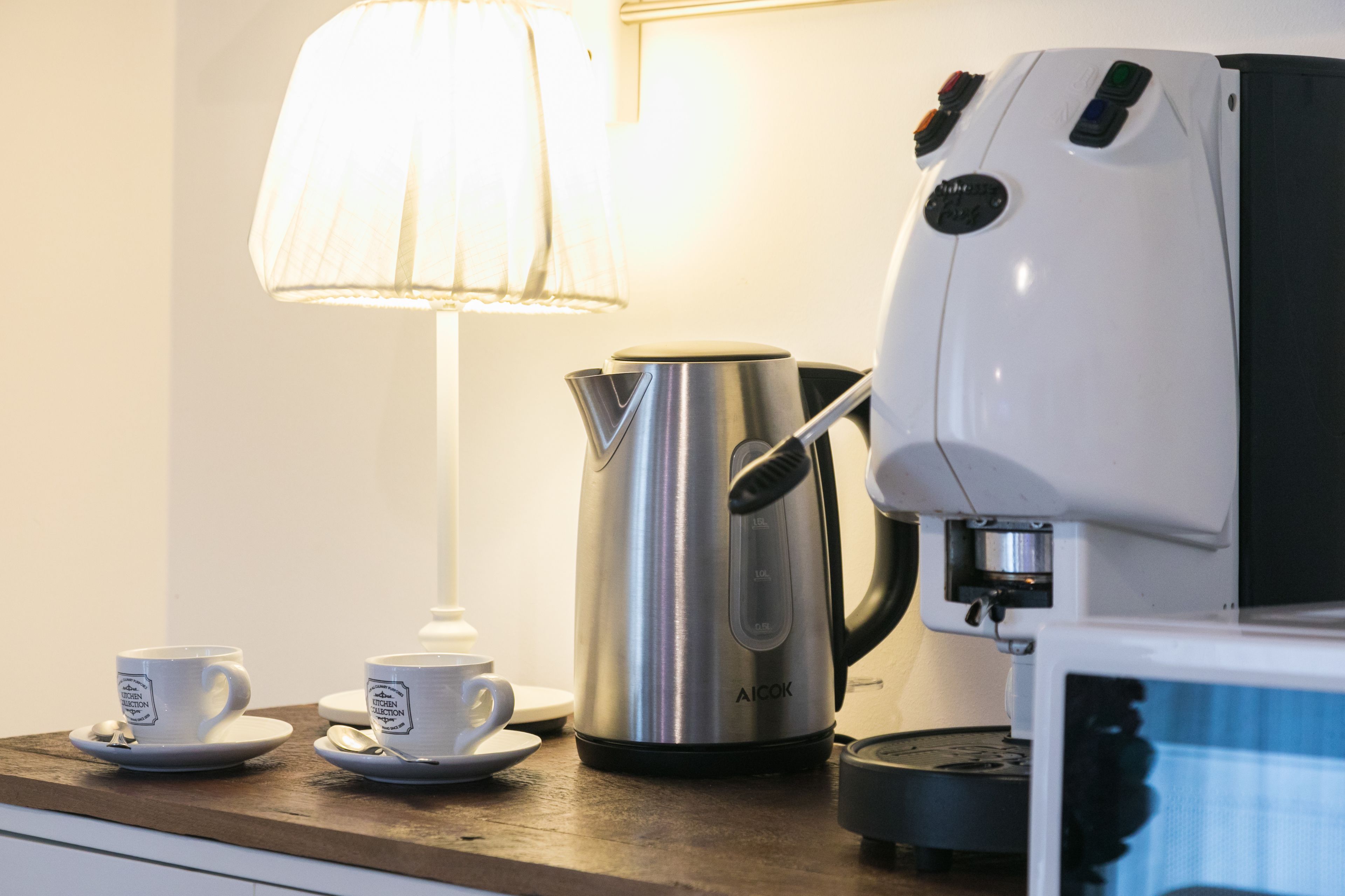 A coffee maker, two cups, a kettle, and a lamp on a wooden table.