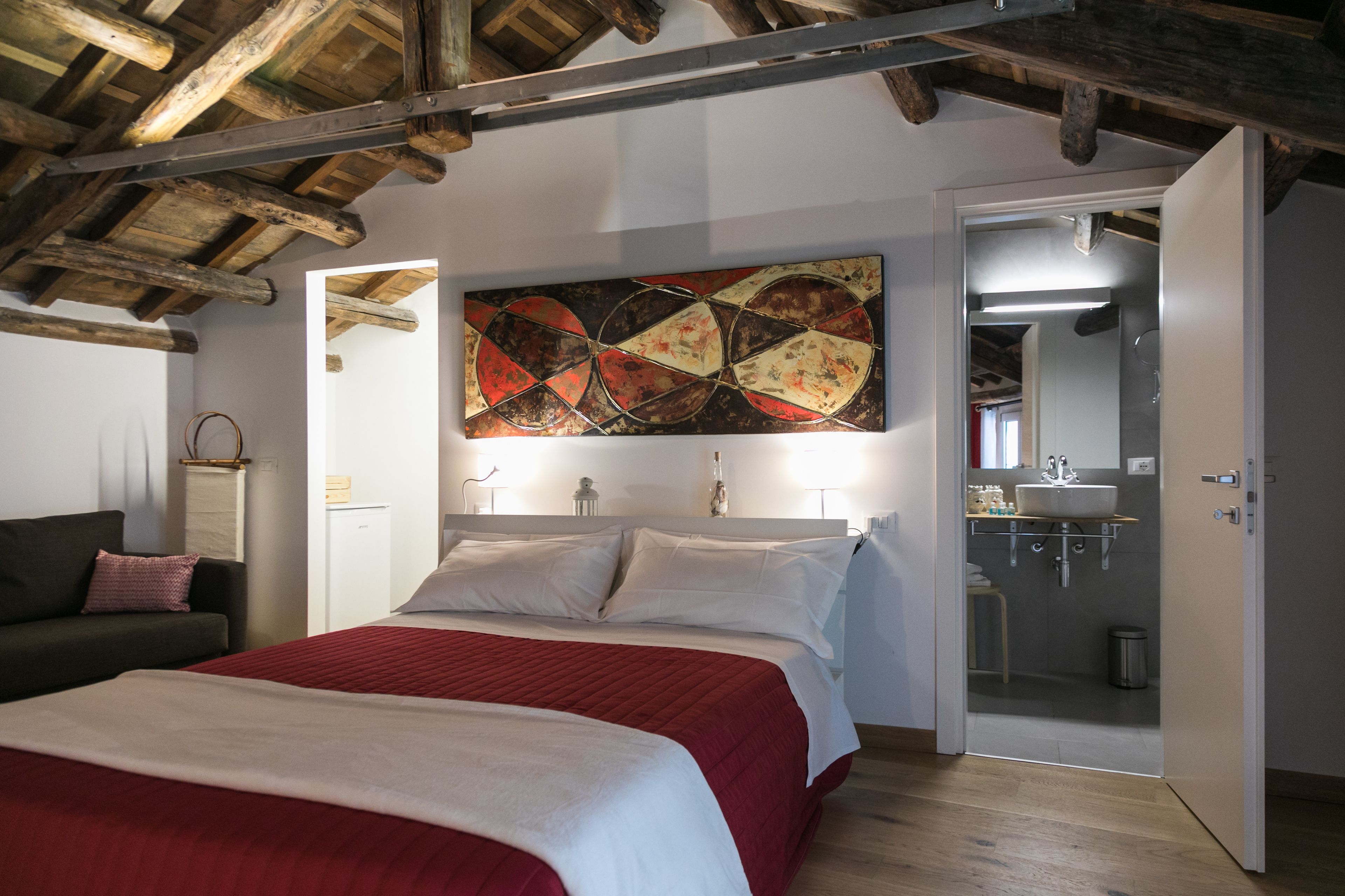 Quarto com teto de madeira, uma cama com colcha vermelha e branca, um quadro abstrato grande na parede e um banheiro visível por uma porta aberta.
