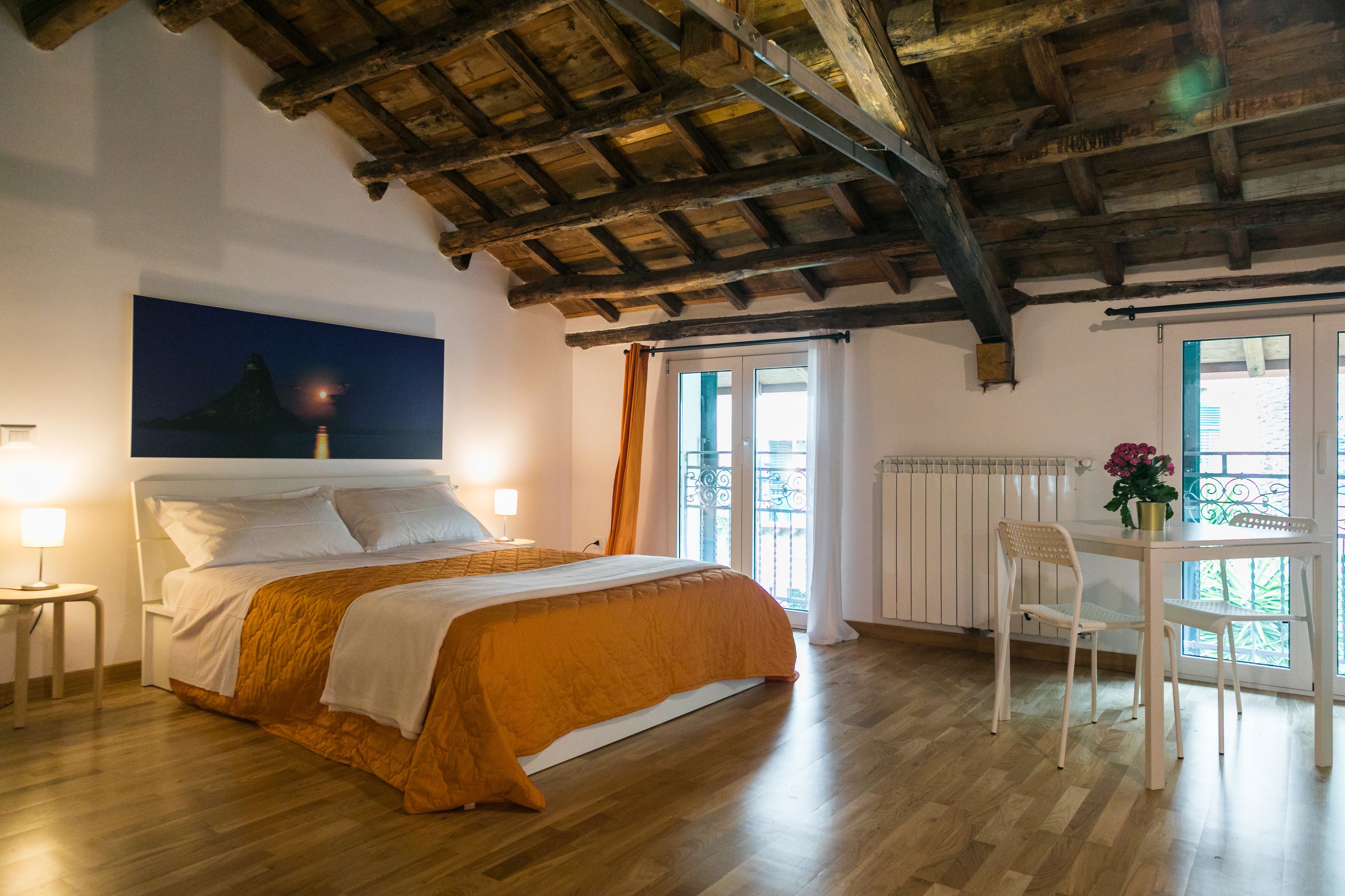 A bedroom with a wooden ceiling, a bed with an orange bedspread, a nightstand, a table with a chair, and a window with a view of greenery.
