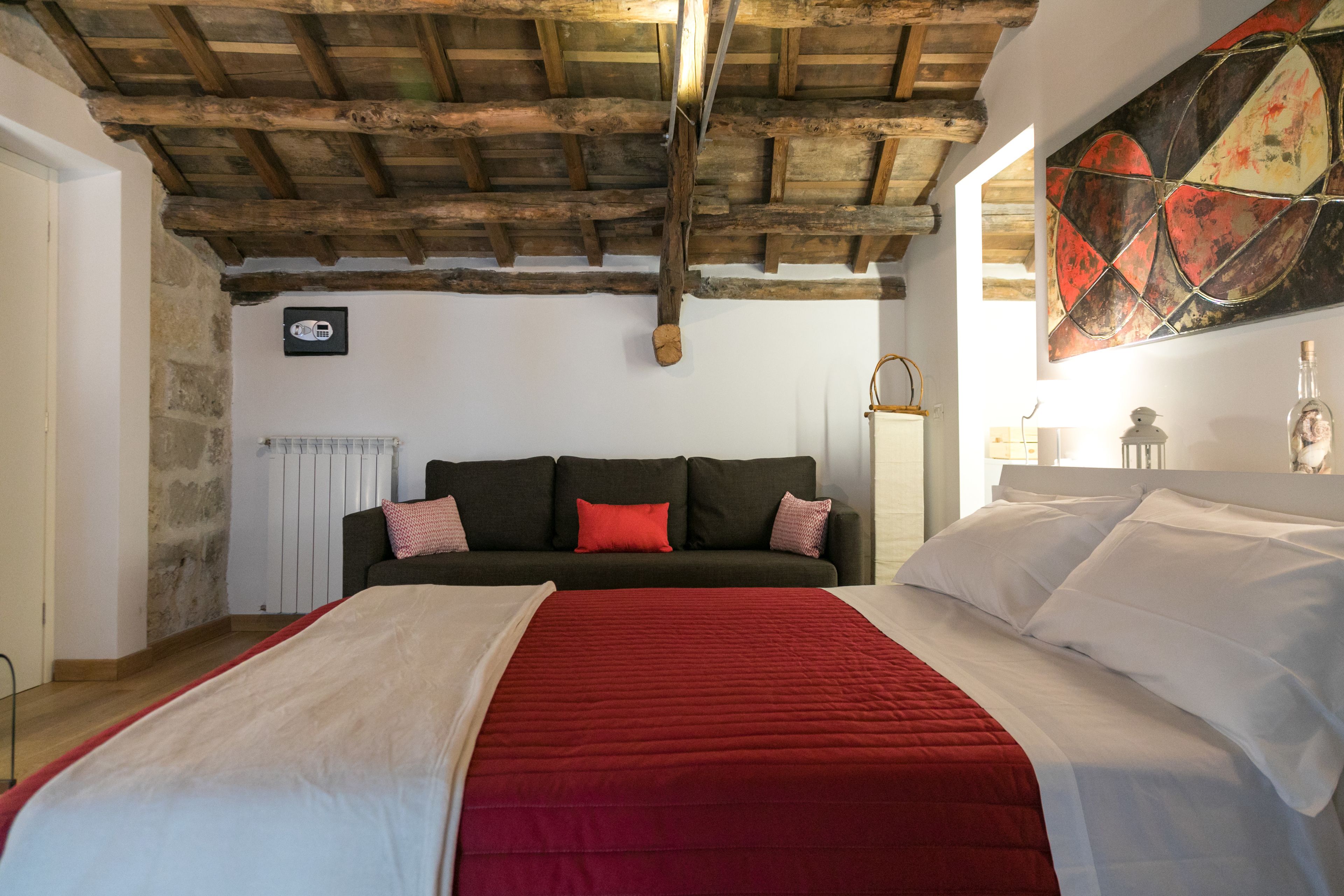 A bedroom with a wooden ceiling, a bed with a red and white bedspread, a dark green sofa with red cushions, and a painting on the wall.
