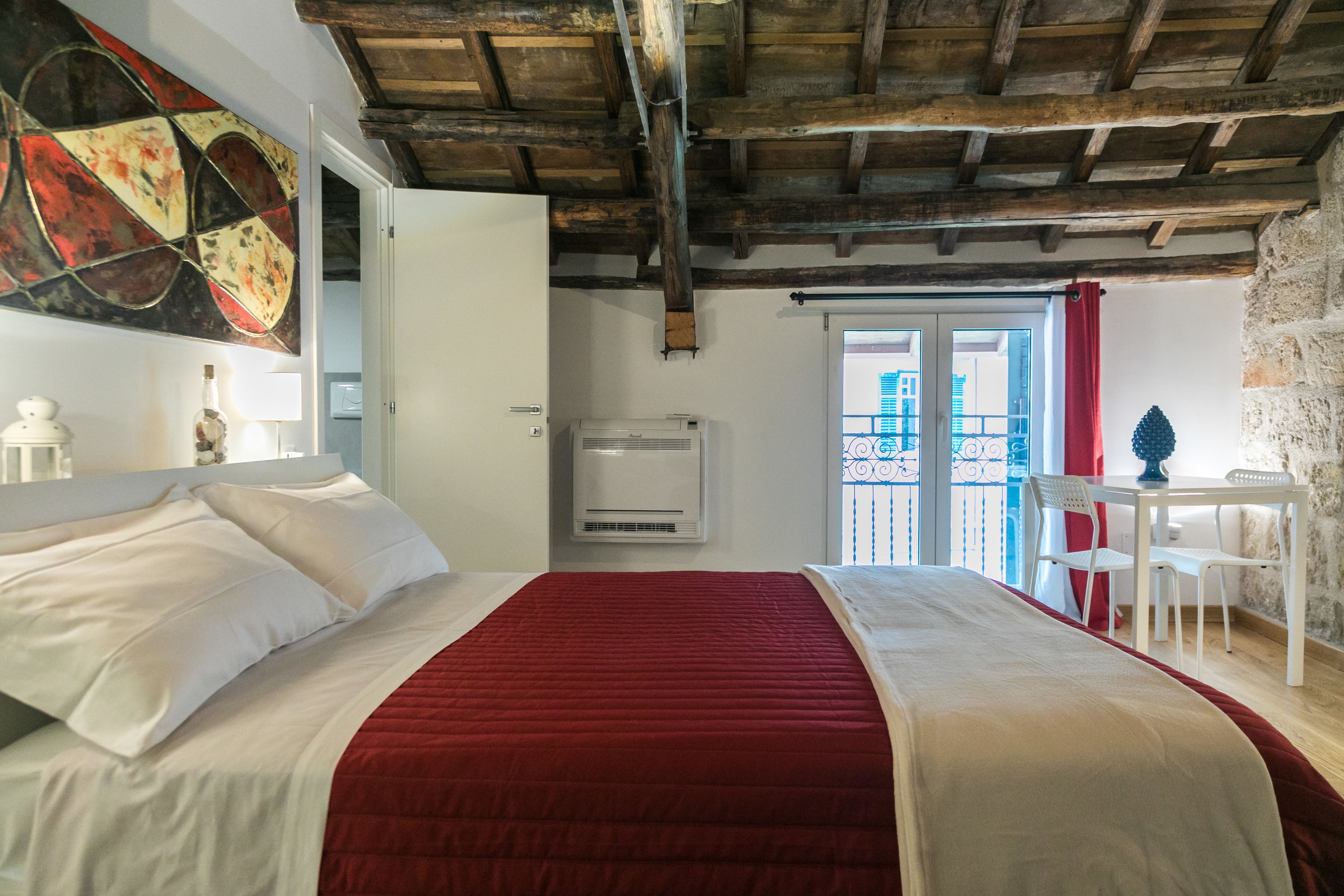 A bedroom with a large bed, a red blanket, a white headboard, a ceiling with exposed beams, a window with a view, and a small table with a chair.