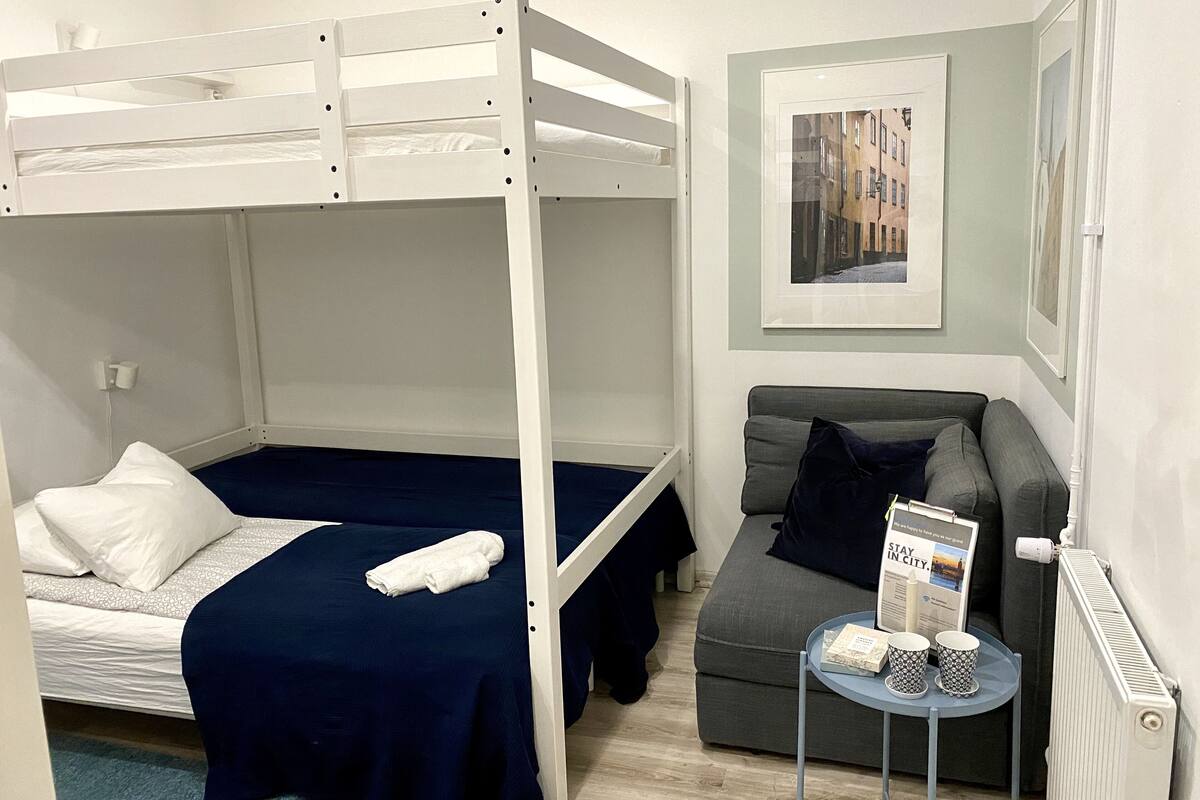 A bunk bed with a blue bedspread and white pillows, a grey sofa with a dark cushion, and a small table with a magazine and cups.