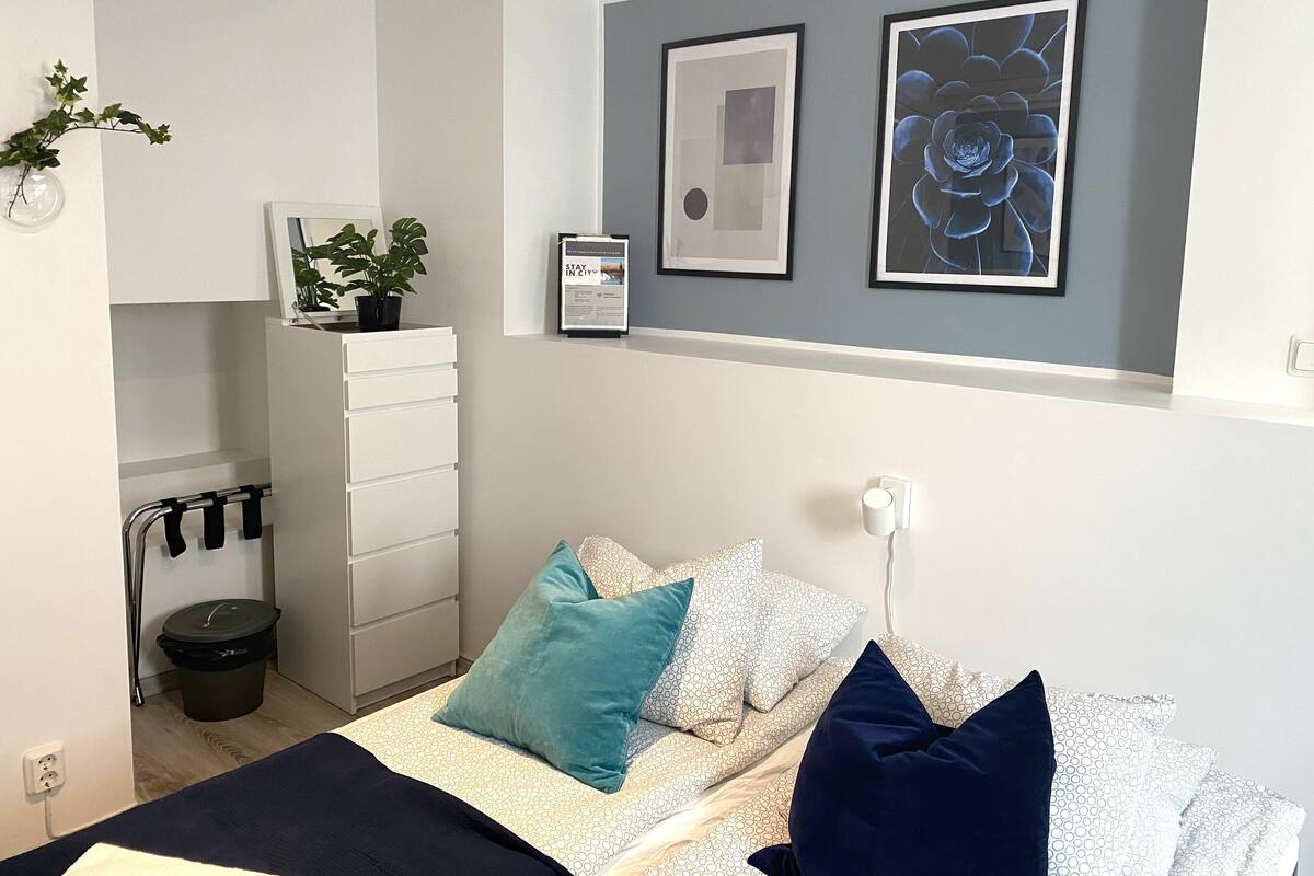 A modern bedroom with a bed, a dresser, a trash bin, and two framed artworks on the wall.