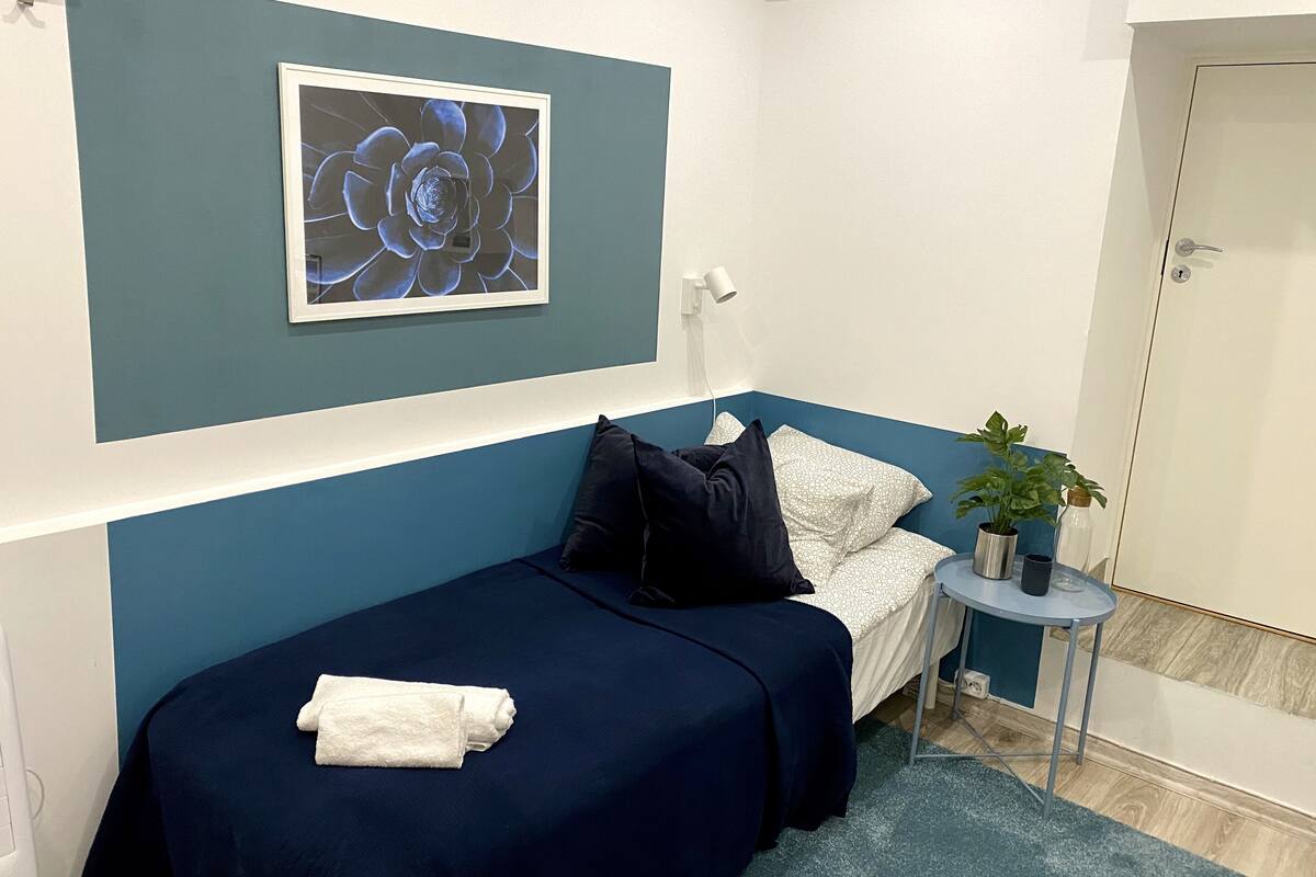A small, neatly arranged room with a bed, a bedside table with a plant, and a framed artwork on the wall.
