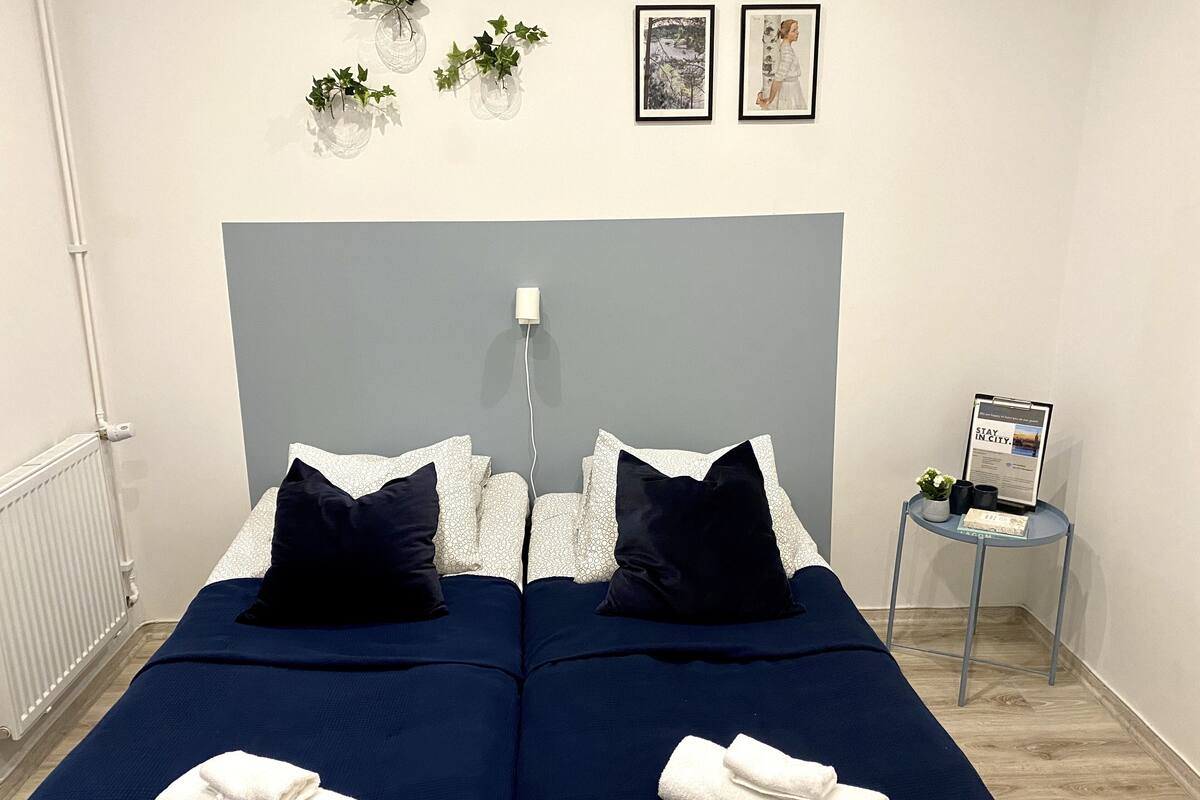 A double bed with two pillows, a small table with a sign, and three wall-mounted plants.