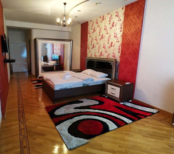 A bedroom with a bed, bedside table, and a mirror. There is a wooden floor and a red and black patterned rug.