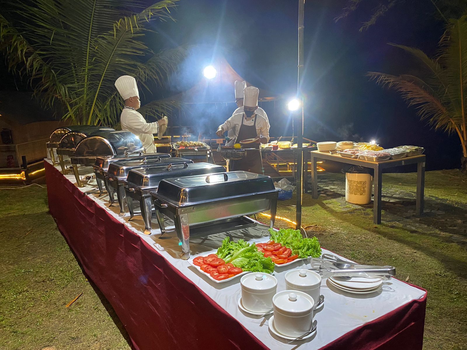 Un dispositiu de buffet amb diverses estacions de menjar, incloent una graella, amanides i plats calents, sota una il·luminació exterior brillant.