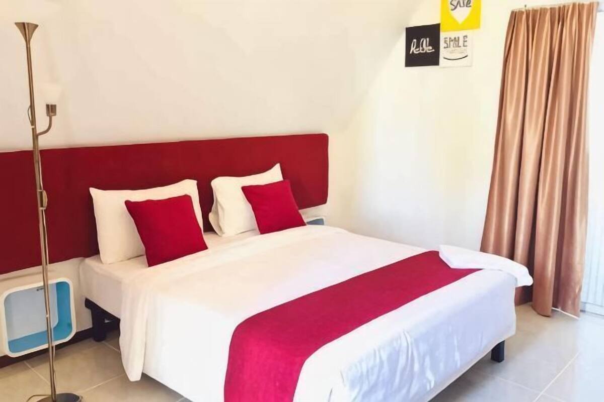Hotellihuone, jossa on sänky, yöpöytä, lattiavalaisin ja verho.