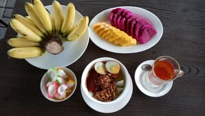 A table with a plate of bananas, a plate of mango slices, a plate of purple fruit, a bowl of egg salad, a bowl of mixed vegetables, and a glass of tea. - D'Bamboo Kamp (Bintan)