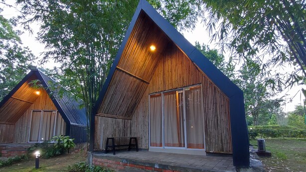A rustic wooden cabin with a sloped roof and large windows, surrounded by lush greenery. - D'Bamboo Kamp (Bintan)