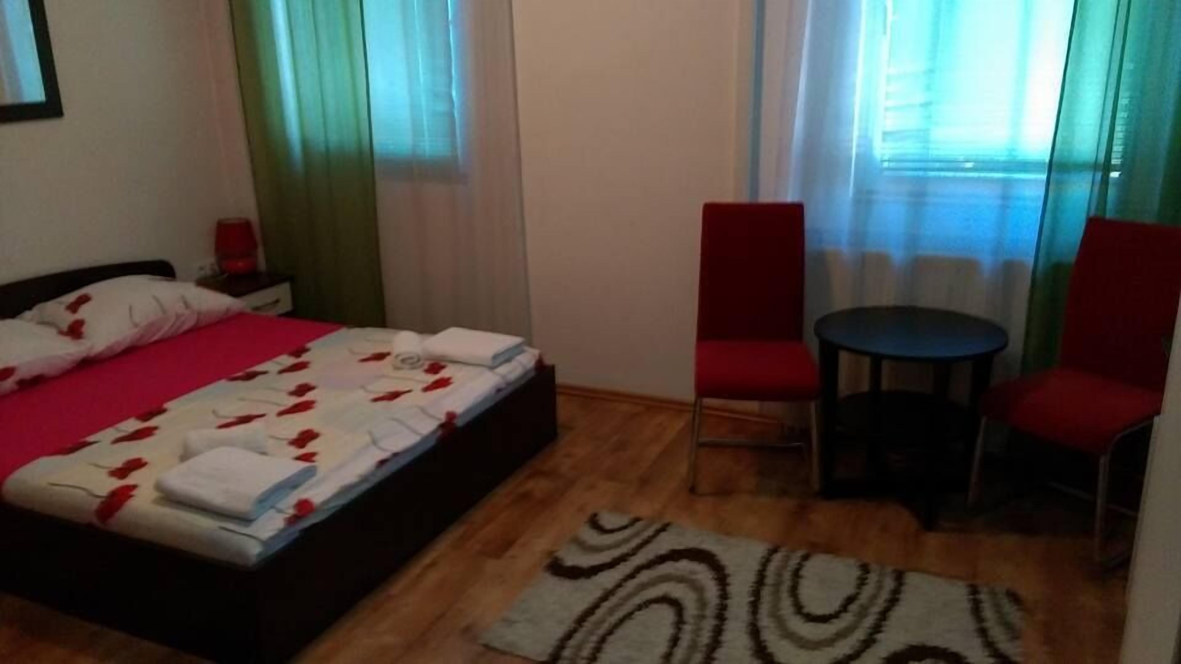 Quarto com uma cama, duas cadeiras vermelhas, uma mesa redonda pequena e um tapete estampado.