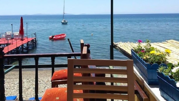 A seaside terrace with a wooden chair, a red table, and a small boat in the water. - Seckin Motel (Ayvacik)