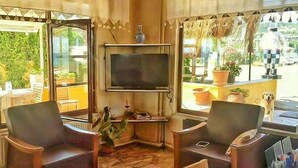 A room with a TV mounted on the wall, two chairs, a small table, and a large window with a view of the outdoors. - Seckin Motel (Ayvacik)