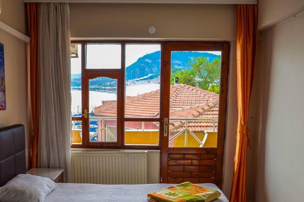 A bedroom with a bed, a window with a view of a town and mountains, and a window with a view of a building with a red roof.