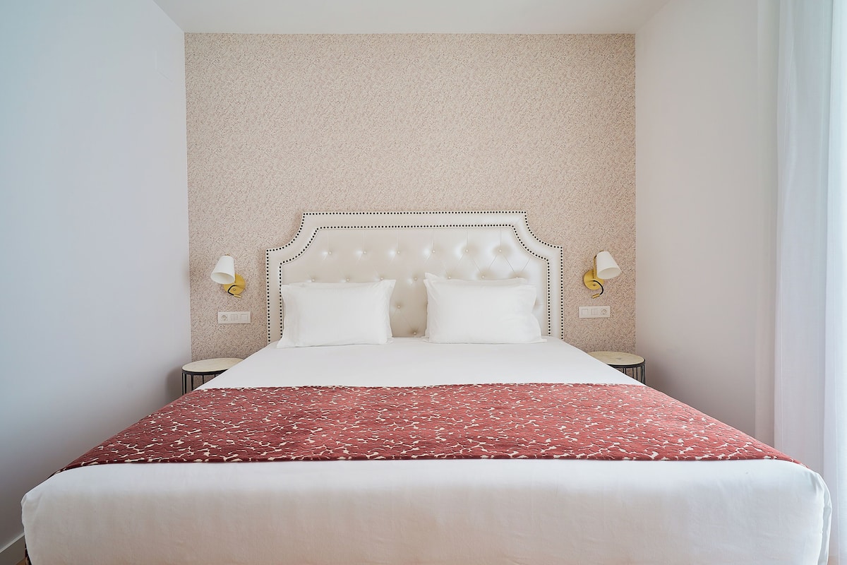 A neatly made bed with a red patterned bedspread, a tufted headboard, and two bedside lamps.