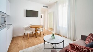 Un salon moderne comprenant une table basse ronde, un canapé, un coin repas avec une table et des chaises, un téléviseur à écran plat fixé au mur et un coin cuisine équipé d’un four à micro-ondes.