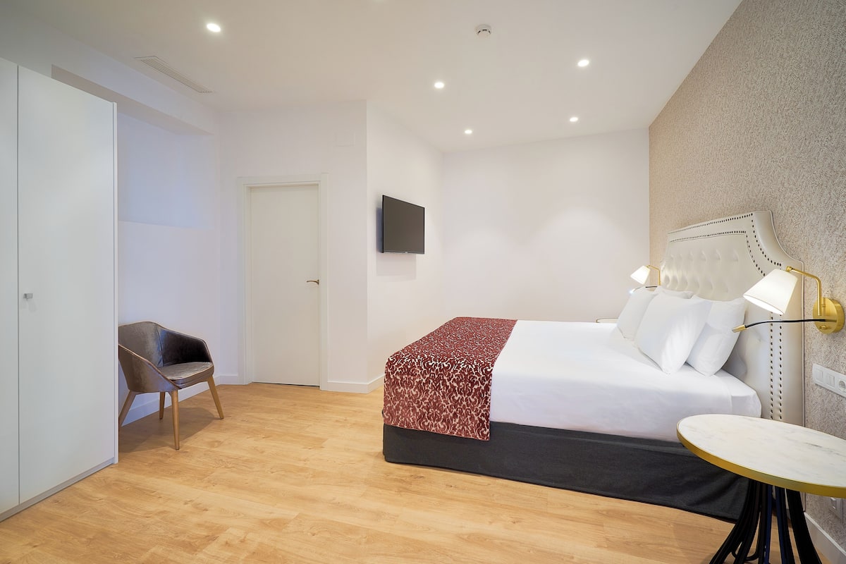 A modern hotel room with a bed, a chair, a small table, and a TV on the wall.