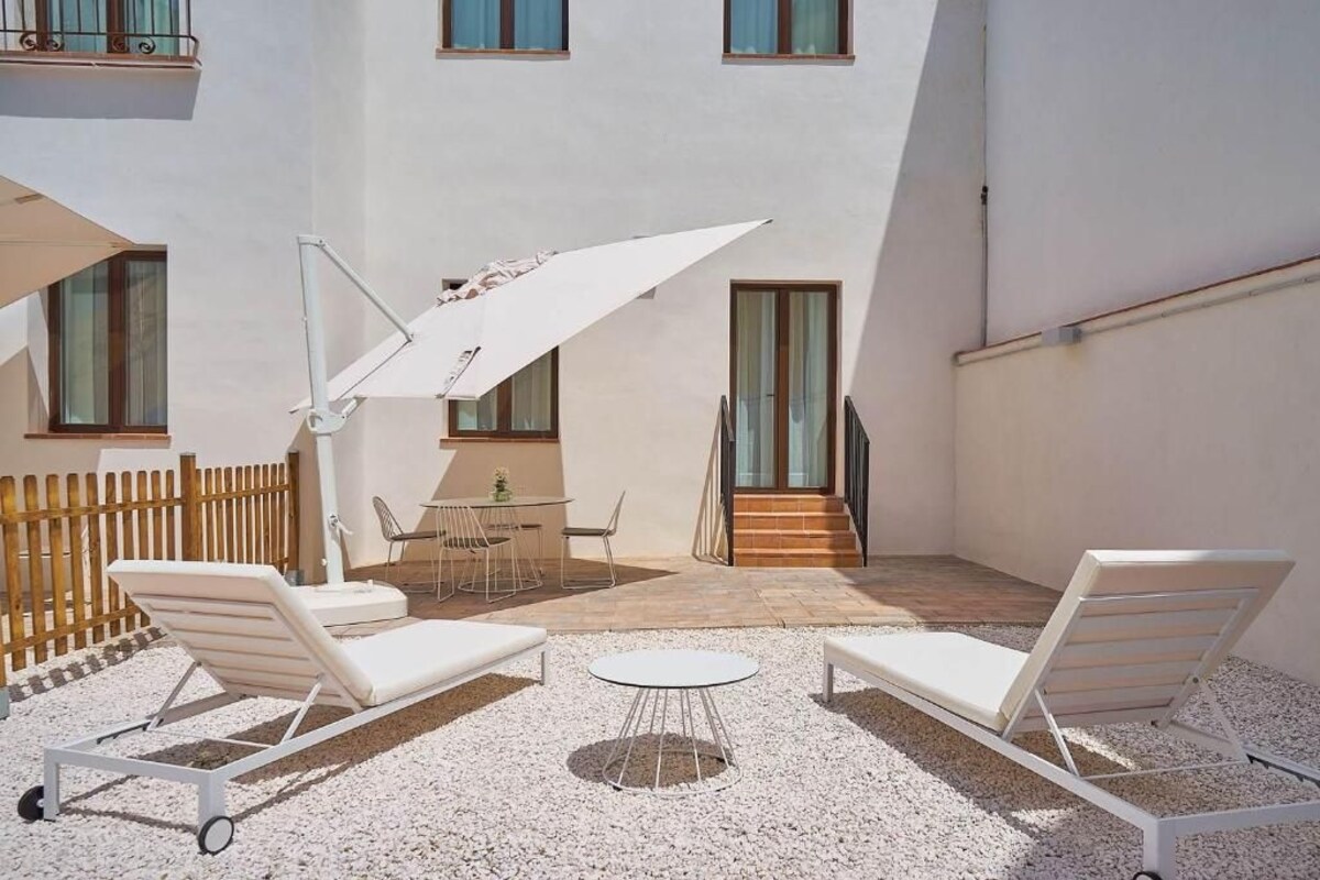 A sunlit outdoor patio with white lounge chairs, a round table, and a large umbrella.