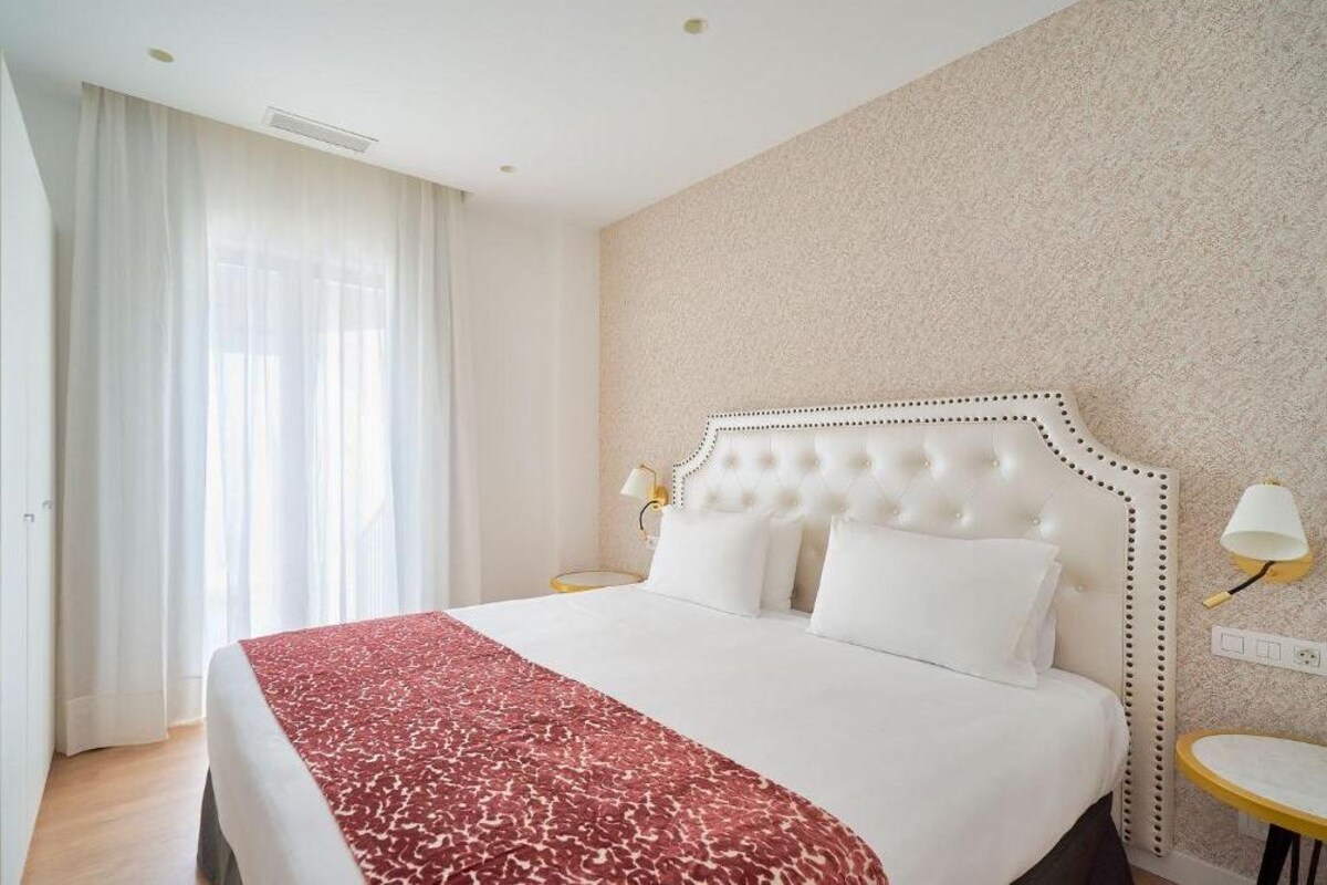 A hotel room with a large bed, white bedding, a red and white patterned throw, a tufted headboard, two bedside lamps, and a small round table.