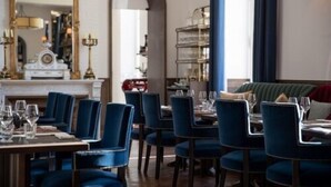 Un comedor con sillas tapizadas en azul y mesas, un espejo grande y una chimenea.
